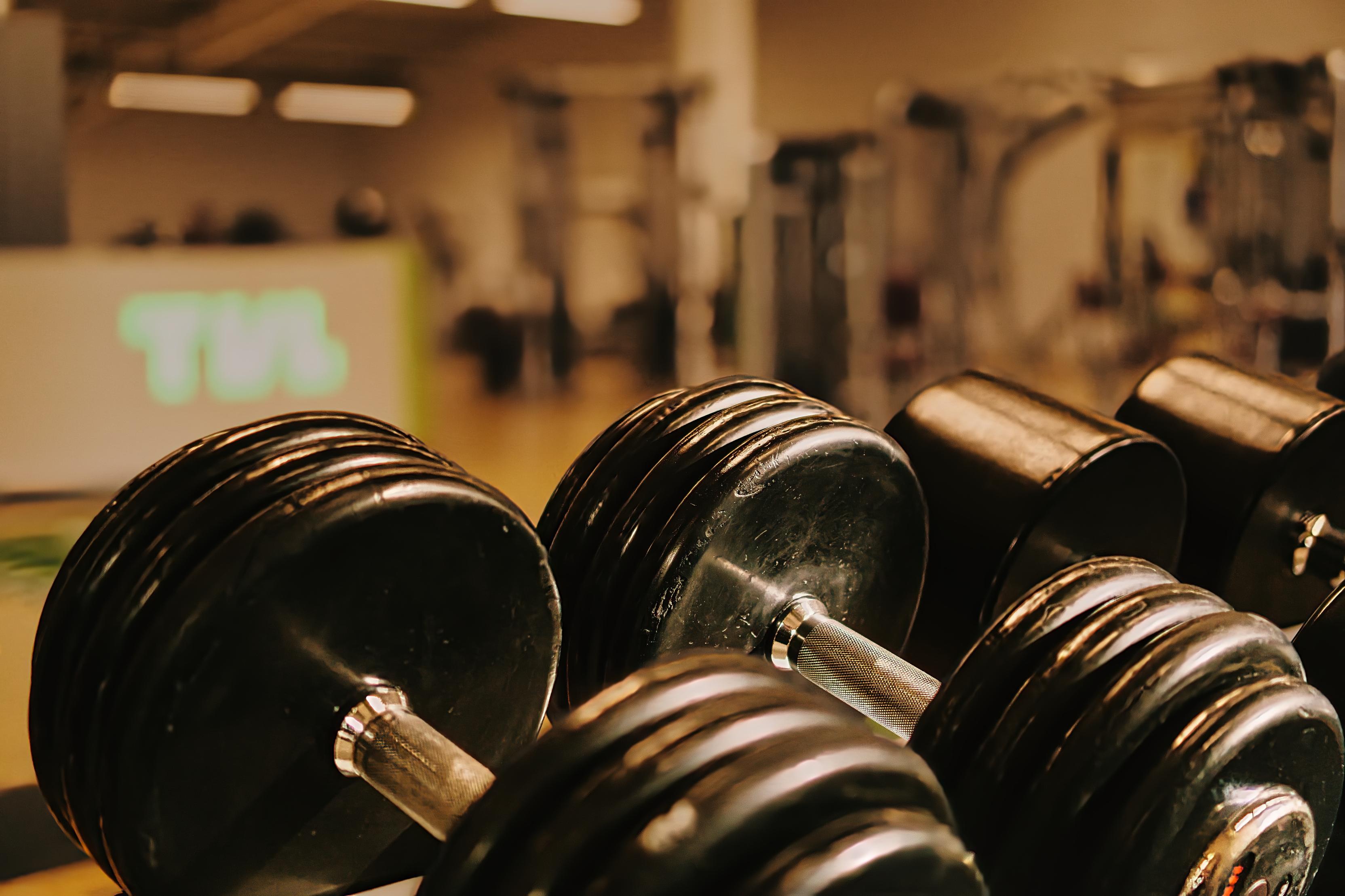 Nahaufnahme mehrerer schwarzer Kurzhanteln auf einem Fitnessstudio-Hintergrund mit Geräten und einer Beleuchtung.